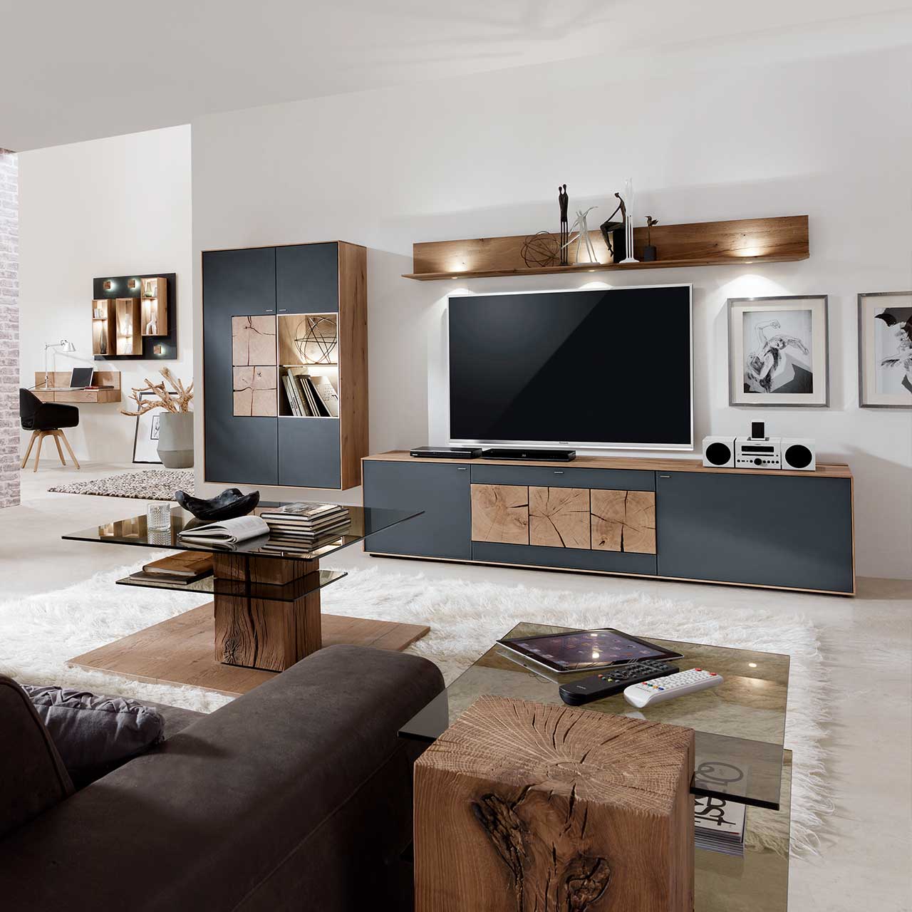 Wohnzimmer mit TV und moderner Wohnwand aus massiver Kerneiche Umato mit Mattglas anthrazit Fronten: Langes Lowboard mit dekorativen Hirnholzscheiben, Hängeregal aus Kerneiche, Hängevitrine mit beleuchtetem Glasausschnitt und Hirnholz.
