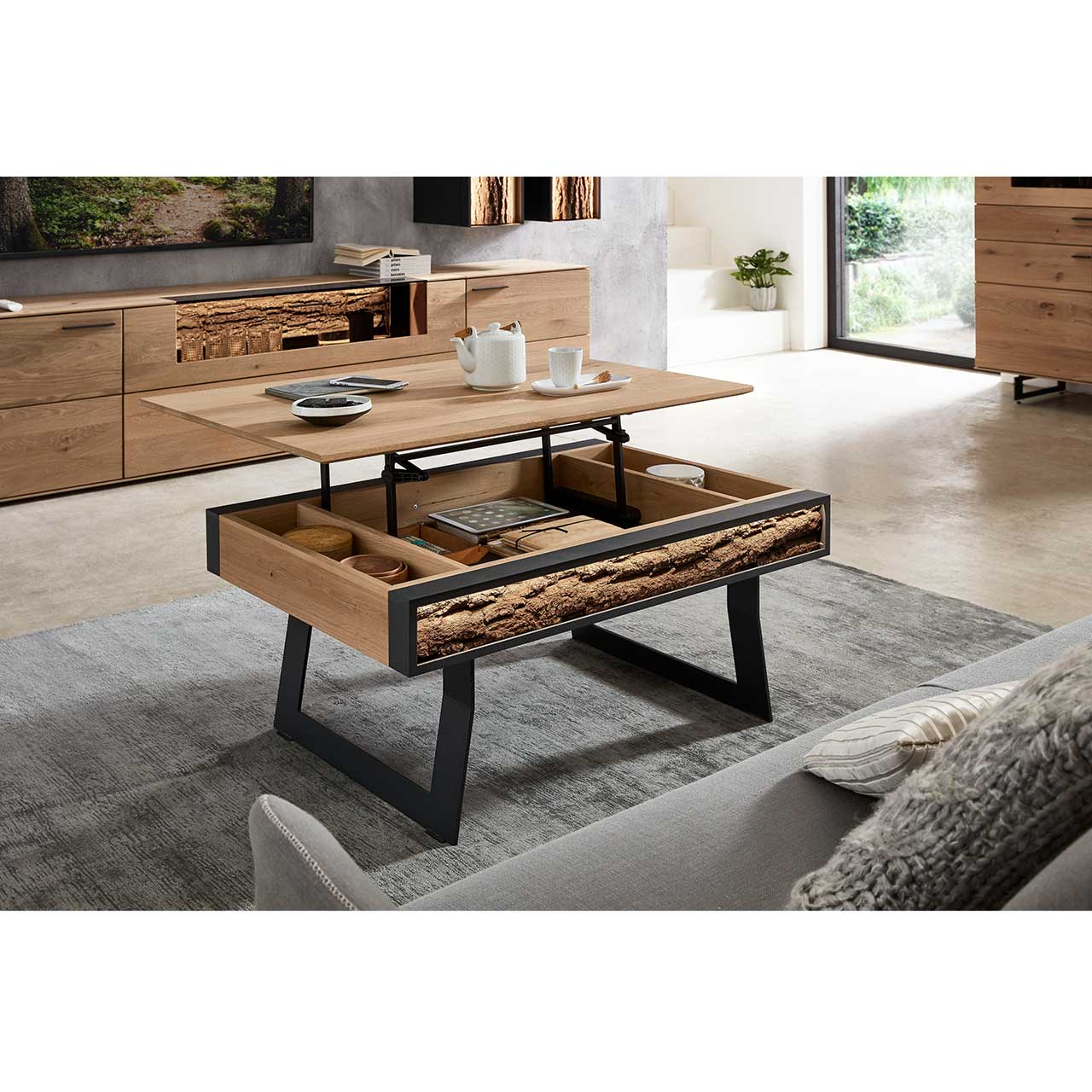 Milieubild Wohnzimmer - Hartmann NATURWERKE - Couchtisch 8400C-1410 - offen Im Wohnzimmer stehender Couchtisch mit hochgefahrener Tischplatte aus Kerneiche, geöffnetes Stauraumfach mit seitlicher Rindenapplikation – auf zwei Metallkufen in Anthrazit.