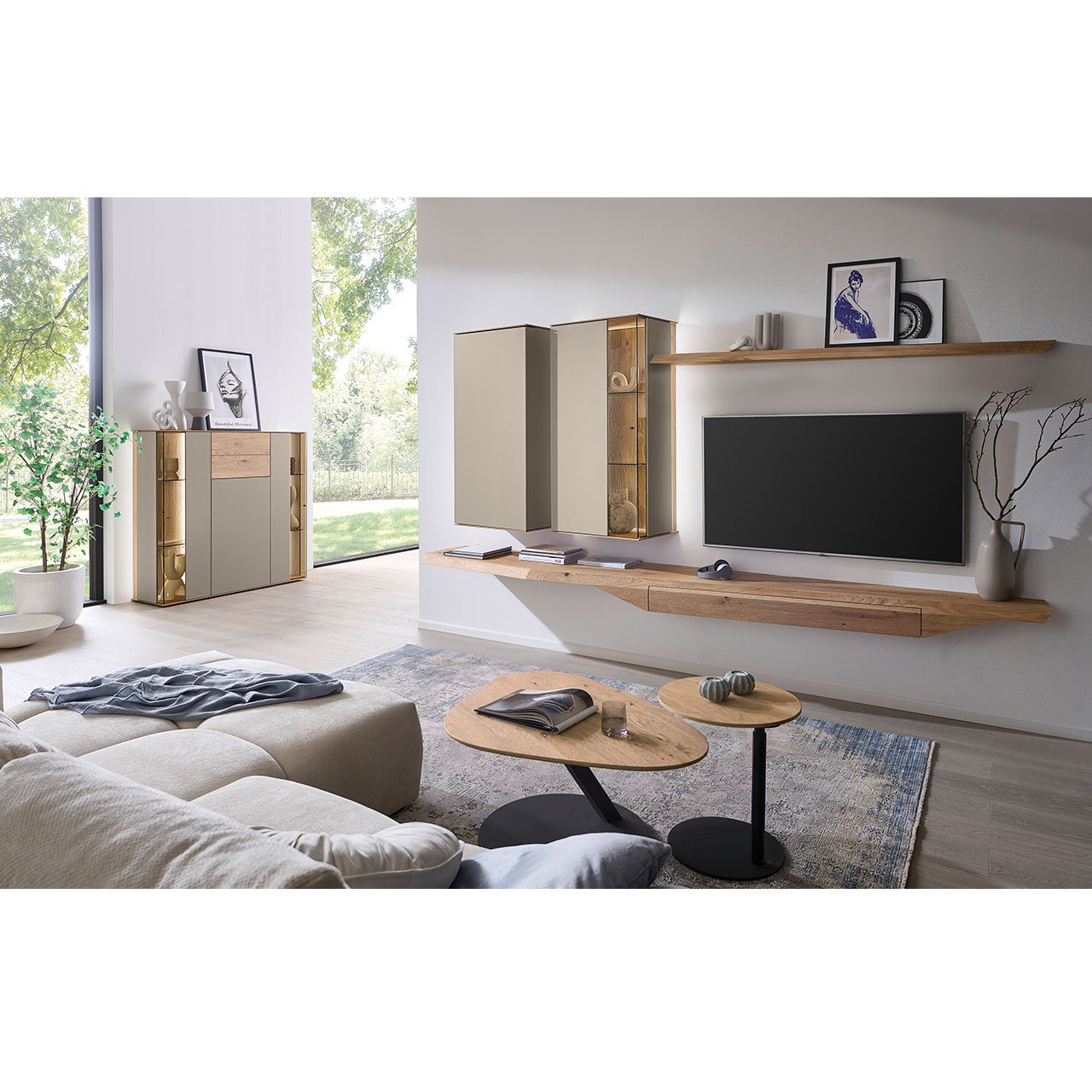 Hartmann Möbel SVEA - Wohnwand 8660 Nr. V26 - vierteilig aus Kerneiche Natur, Lack Sand - Milieubild Wohnzimmer mit moderner Massivholzwohnwand bestehend aus Hängelowboard mit Schubkasten, Hängevitrine, Hängeschrank sowie Wandboard - im asymmetrischen Design. Links passendes Sideboard mit Glastüren, davor zwei Beistelltische aus Holz.