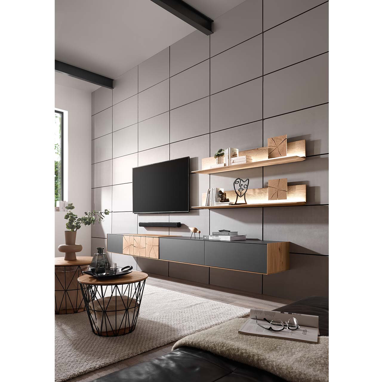 Wohnzimmer mit TV und moderner Wohnwand aus massiver Kerneiche Umato mit Mattglas anthrazit Fronten, mit langen Hängelowboard und zwei Wandpaneelen mit Hirnholzscheiben - an gefliester grauer Wand. Im Vordergrund zwei runde Couchtische mit Korbgestell.