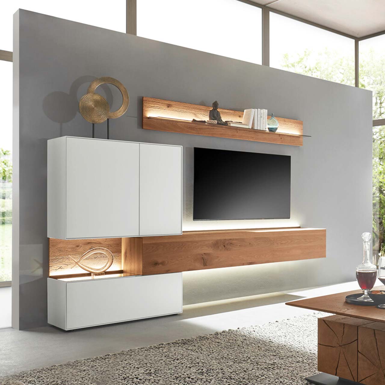 Moderne Wohnwand aus massiver Birke weiß lackiert mit Kerneiche Umato bestehend aus einem Holz-Hängelowboard seitlich eingefasst in einen weißen Unter,- und Hängeschrank mit beleuchteter Rückwand und einem Holz-Wandpaneel mit Glasablageboden.
