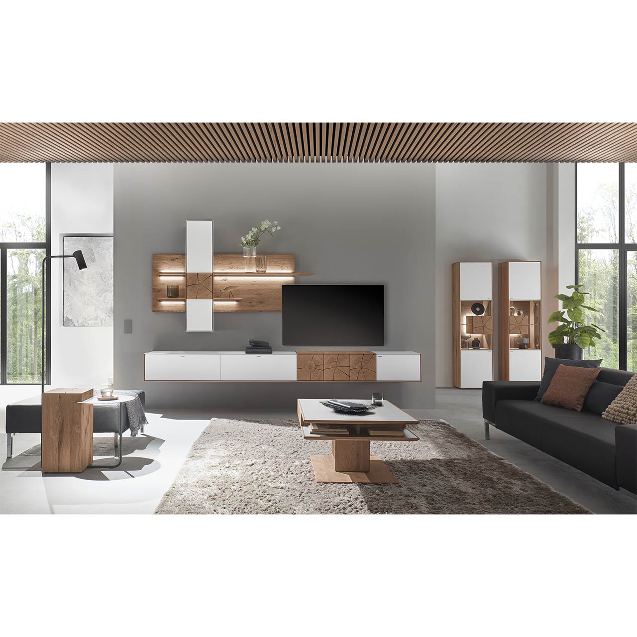 Moderne Wohnwand aus massiver Kerneiche mit Mattglas weißen Fronten, mit Hängelowboard, Wandpaneel mit Regalböden und Hängeschrank. Rechts im Hintergrund zwei passende Standvitrinen. Im Vordergrund ein Holz-Couchtisch mit Ablage.