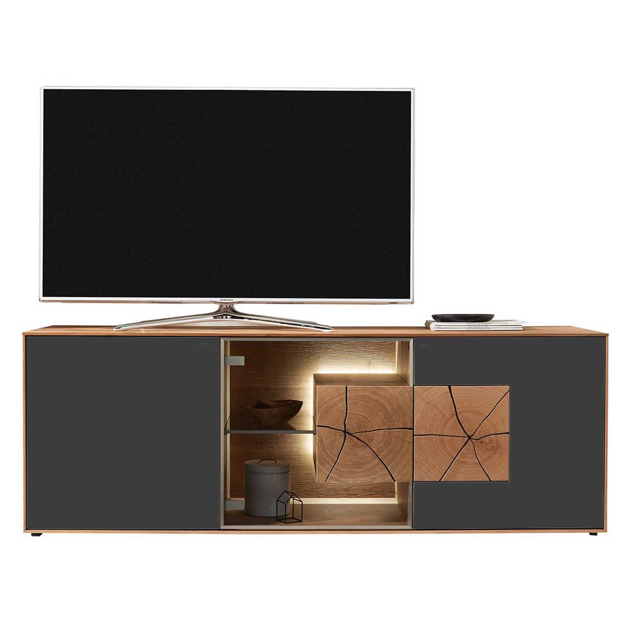Hartmann CAYA - Lowboard 7170-3171A Großer TV platziert auf modernem Lowboard aus massiver Kerneiche Umato mit Fronten aus Mattglas anthrazit, mittig Glastür mit Glasboden und beleuchtetem Hirnholzblock.