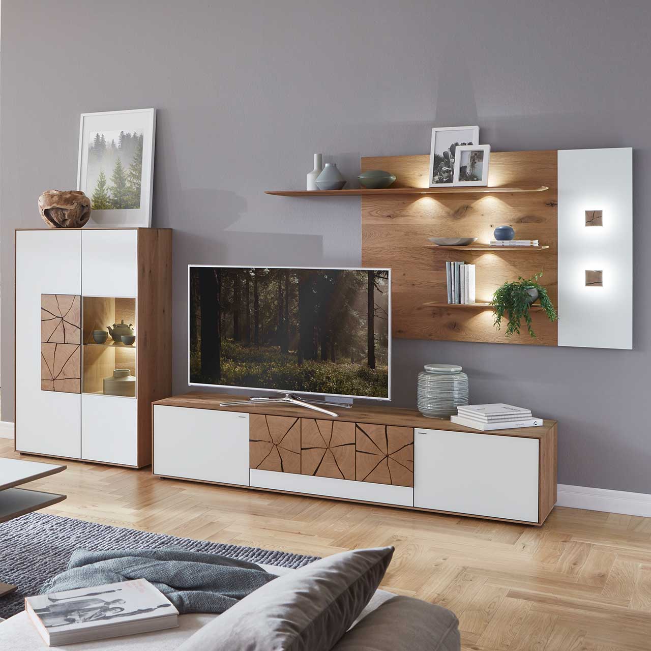 Wohnzimmer mit moderner dreiteiliger Massivholz Wohnwand mit Mattglas weiß Fronten und dekorativen Hirnholzscheiben: Langes Lowboard mit Hirnholz, Wandpaneel mit Regalböden, Highboard mit beleuchtetem Glasausschnitt und Hirnholz.