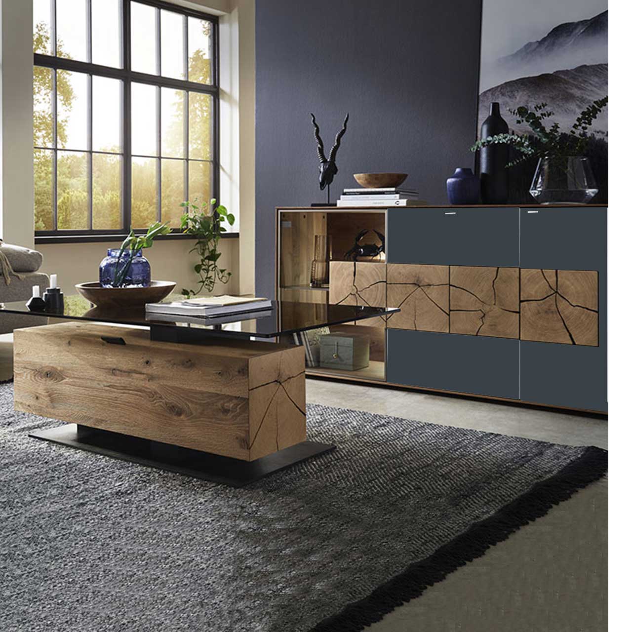 Bildausschnitt eines Wohnzimmers mit modernem Lowboard aus massiver Kerneiche Umato mit anthrazitfarbenen Mattglasfronten, drei rechteckigen Hirnholzscheiben und Glastür mit beleuchtetem Glasboden und Hirnholzblock, davor passender Couchtisch.