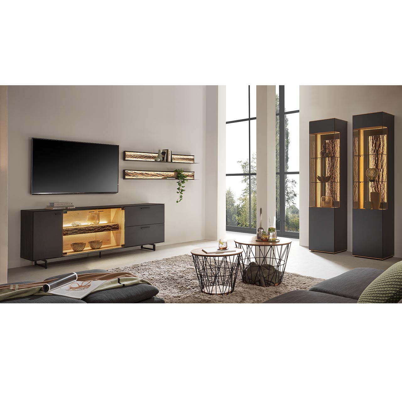 Modernes Wohnzimmer mit Wohnwand bestehend aus einem Lowboard aus massiver Kerneiche Natur, anthrazit gebeizt mit beleuchtetem Glasausschnitt auf Metallkufen und zwei Wandregale aus Metall mit sichtbarer Baumrinde.Rechts zwei Standvitrinen.
