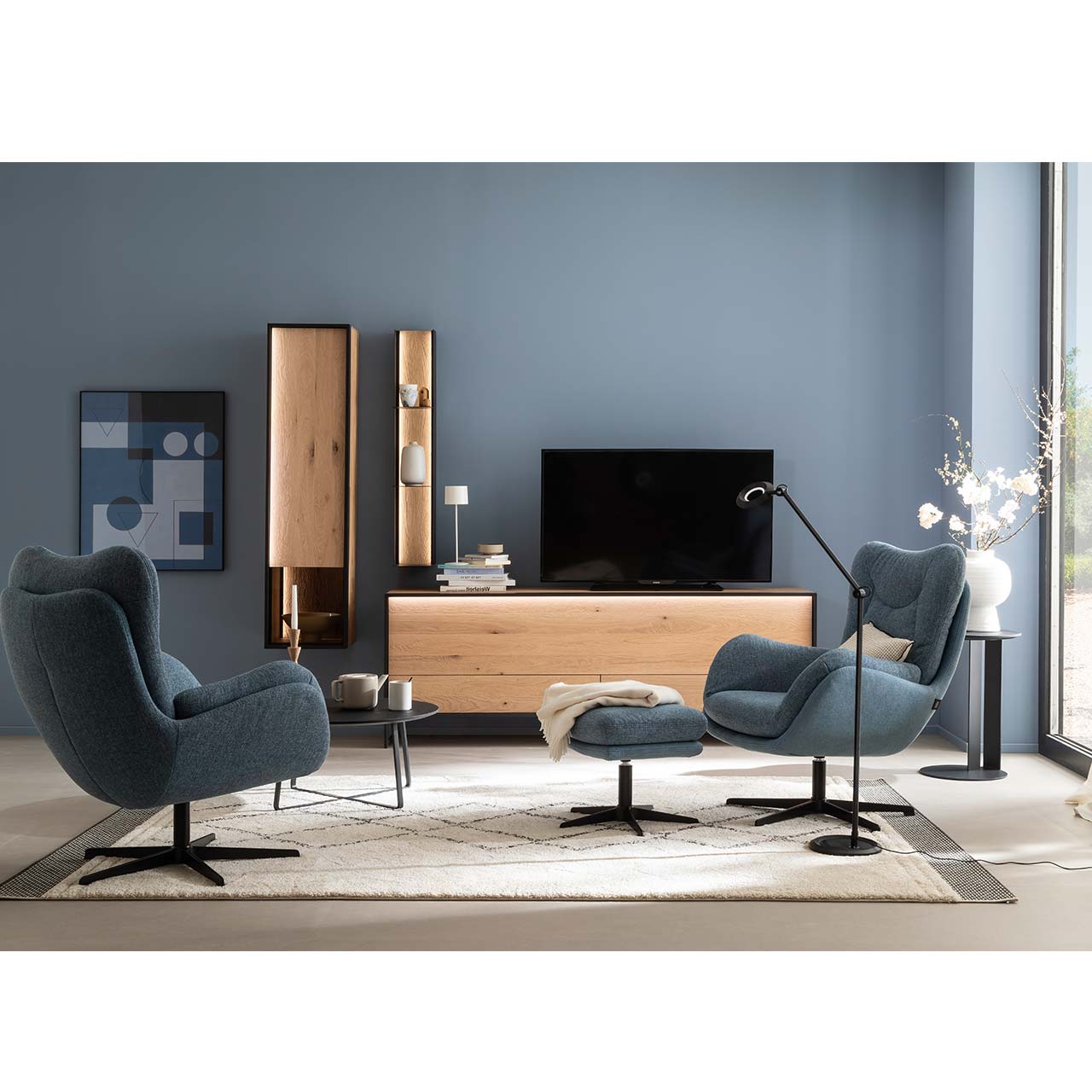Eingerichtetes Wohnzimmer mit blauen Wänden und einer dreiteiligen Wohnwand aus massiver Kerneiche Natur bestehend aus einem schlichten Lowboard auf Metallkufen, einem Hängeschrank mit Glasausschnitt und einem Metallregal mit Riffholz.