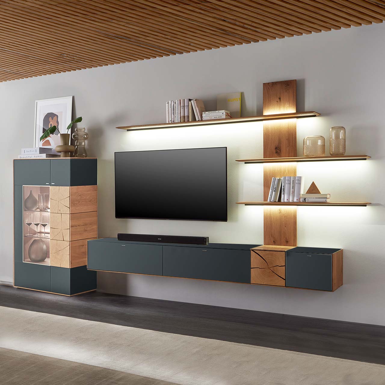 Massivholz Wohnwand aus Kerneiche Umato mit Mattglas anthrazit Fronten. Unten ein Hängelowboard mit dekorativer Holzapplikation, seitlich ein Wandpaneel mit beleuchteten Regalböden, links passende Standvitrine, dazwischen hängender Fernseher.