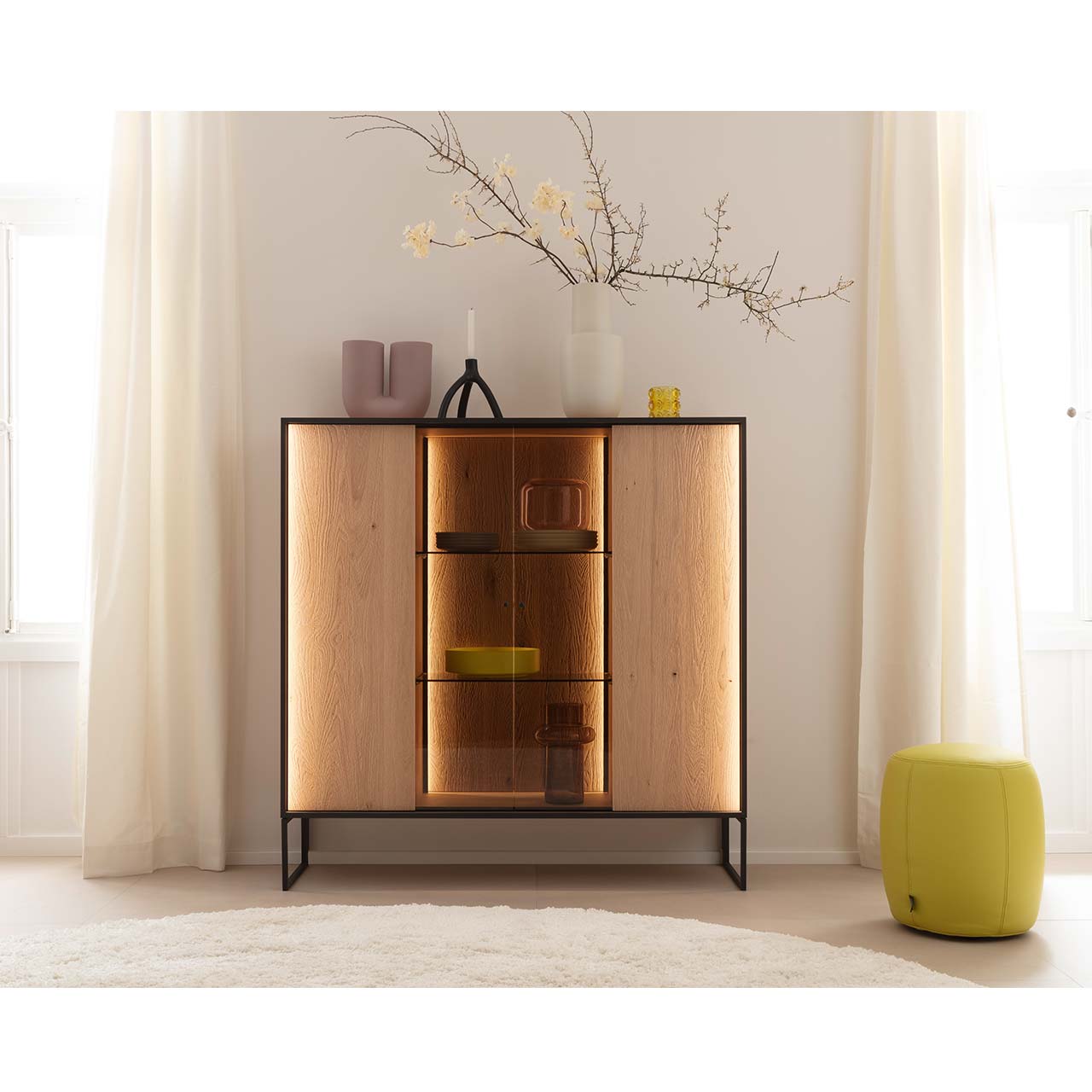 Modernes Highboard aus massiver, sandgestrahlter Kerneiche Natur mit beleuchtetem, rundum Metallrahmen und zwei filigranen Metallfüßen in Anthrazit. Mit zwei Holztüren links und rechts, mittig verglaster Vitrinenbereich mit beleuchteten Glaseinlegeböden. 