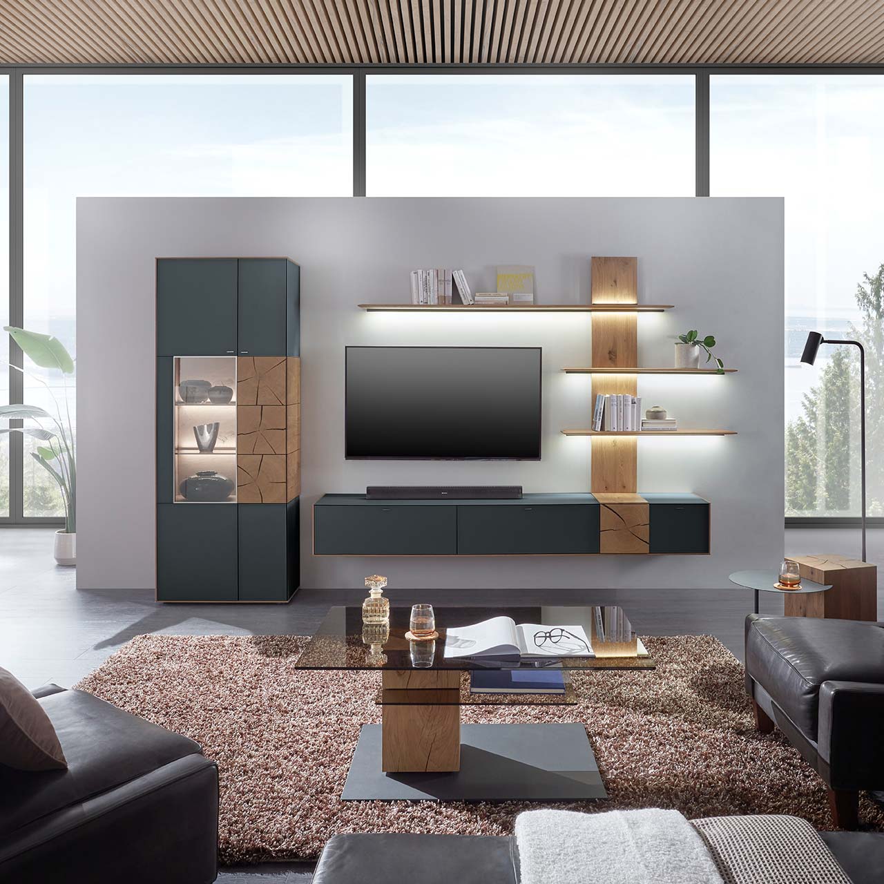 Moderne Wohnwand aus massiver Kerneiche mit anthrazitfarbenen Fronten, mit Hängelowboard, Wandpaneel mit Regalböden und Standvitrine. Im Vordergrund der passende Couchtisch mit rechteckiger Glasplatte.