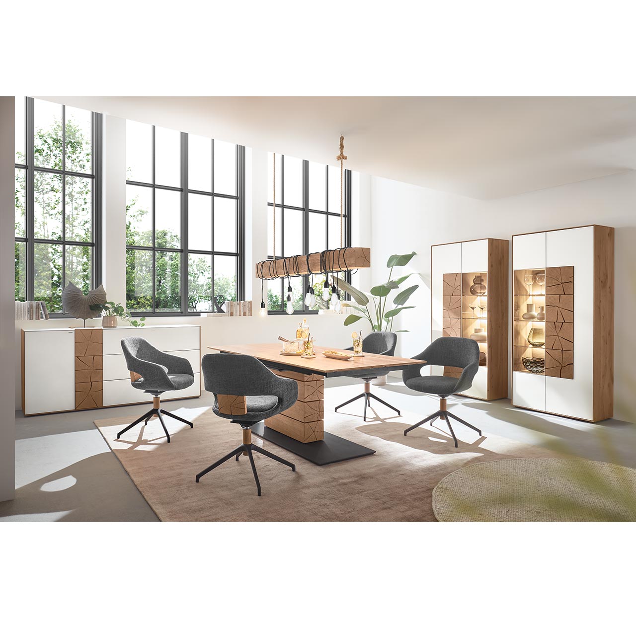 Esszimmer mit rechteckigem Holztisch und Säule aus massiver Kerneiche und vier modernen Stühlen in dunkelgrauem Stoff mit Holz-/Metallgestell. Darüber hängend eine Balkenleuchte. Rechts an der Wand zwei passende Standvitrinen.