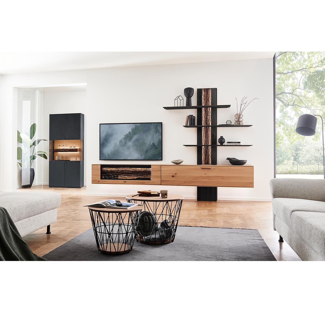 Wohnzimmer mit Wohnwand aus zwei Hängelowboards in massiver Kerneiche Natur, teils verglast, einem Wandpaneel mit beleuchteter Rinden-Rückwand und drei langen Ablageböden, dazu links eine Standvitrine in anthrazit gebeizter Kerneiche.