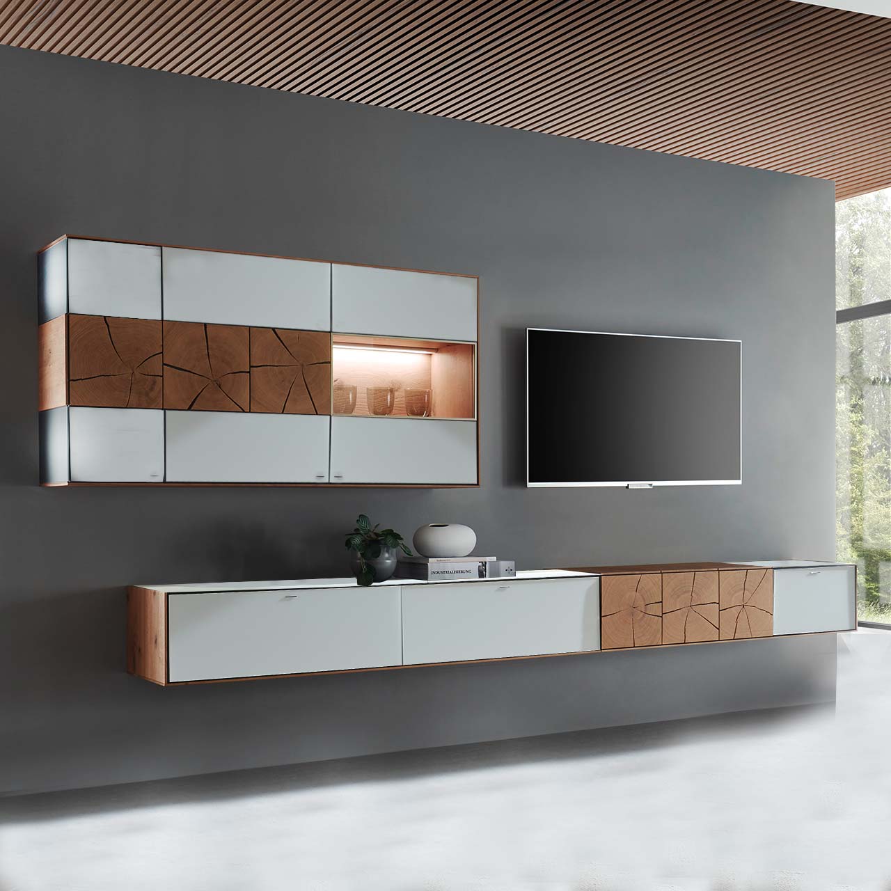 Hartmann CAYA - Wohnwand 7170 Nr. V82W - zweiteilig Moderne zweiteilige Massivholz Wohnwand aus Kerneiche mit Mattglas anthrazit Fronten. Unten ein langes, schmales Hängelowboard mit dekorativen Hirnholzscheiben, darüber die passende, beleuchtete Hängevitrine. Rechts daneben hängender Fernseher.