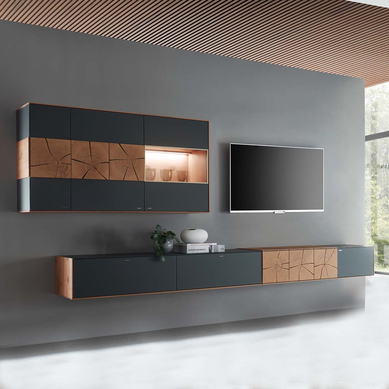 Moderne zweiteilige Massivholz Wohnwand aus Kerneiche mit Mattglas anthrazit Fronten. Unten ein langes, schmales Hängelowboard mit dekorativen Hirnholzscheiben, darüber die passende, beleuchtete Hängevitrine. Rechts daneben hängender Fernseher.