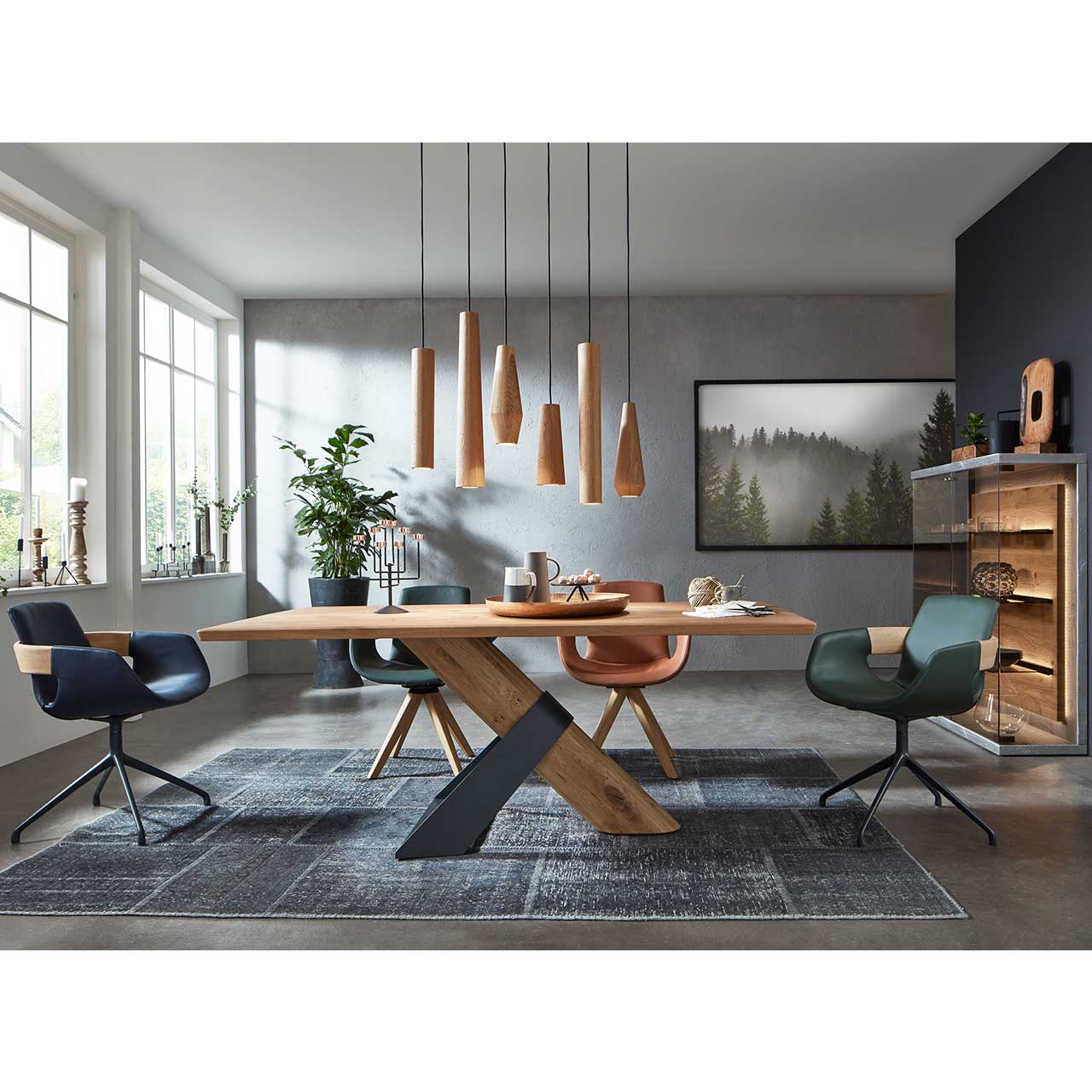 Modernes Esszimmer mit massivem Holztisch, vier Stühlen mit unterschiedlich farbigen Bezügen mit Holz - und Metallgestell. Darüber eine moderne Pendelleuchte mit sechs unterschiedlichen geometrischen Holzkörpern. Rechts modernes Highboard mit Glasfront.