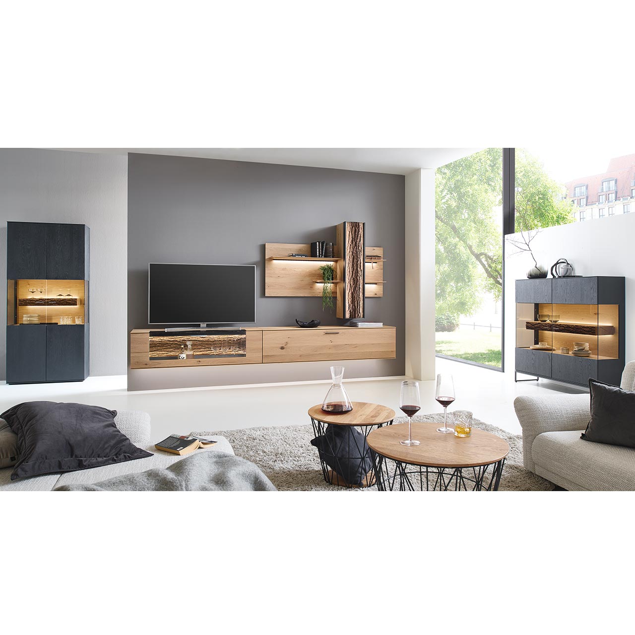 Milieubild Hartmann RUNA 8410 - Wohnwand Nr. V46 - dreiteilig - Kerneiche Natur und Baumrinde Wohnzimmer mit Wohnwand aus zwei Hängelowboards in massiver Kerneiche Natur, teils verglast, einem Wandpaneel mit beleuchteten Böden und Stauraumschrank, dazu links eine Standvitrine und rechts das passende Highboard in anthrazit gebeizter Kerneiche.
