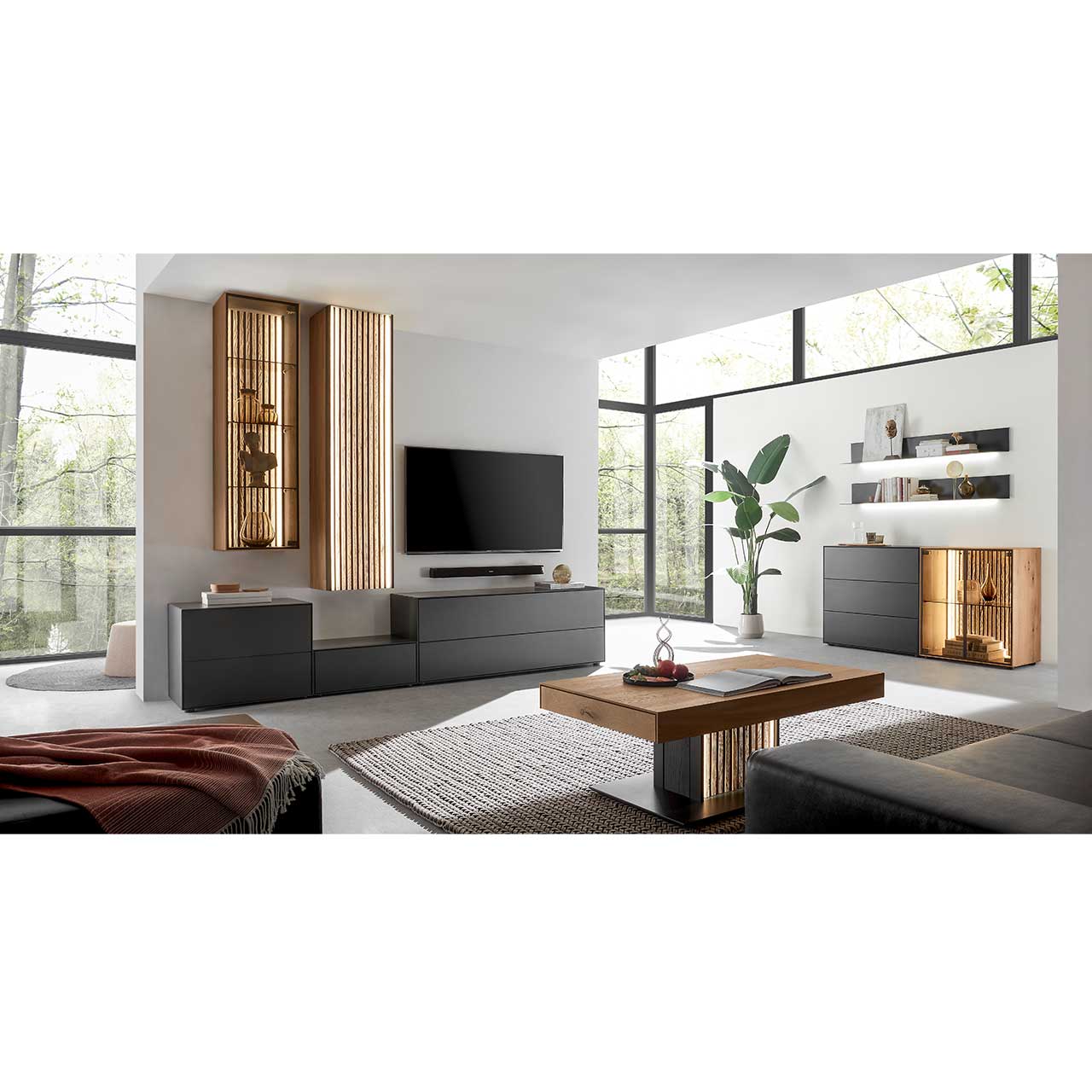 Modern eingerichtetes Wohnzimmer mit fünfteiliger Massivholz-Wohnwand bestehend aus Lowborads in anthrazit, Hängevitrine und Hängeschrank in hellem Holz mit Lamellen. Davor der passende helle Couchtisch. Rechts ein Sideboard und Wandregale.