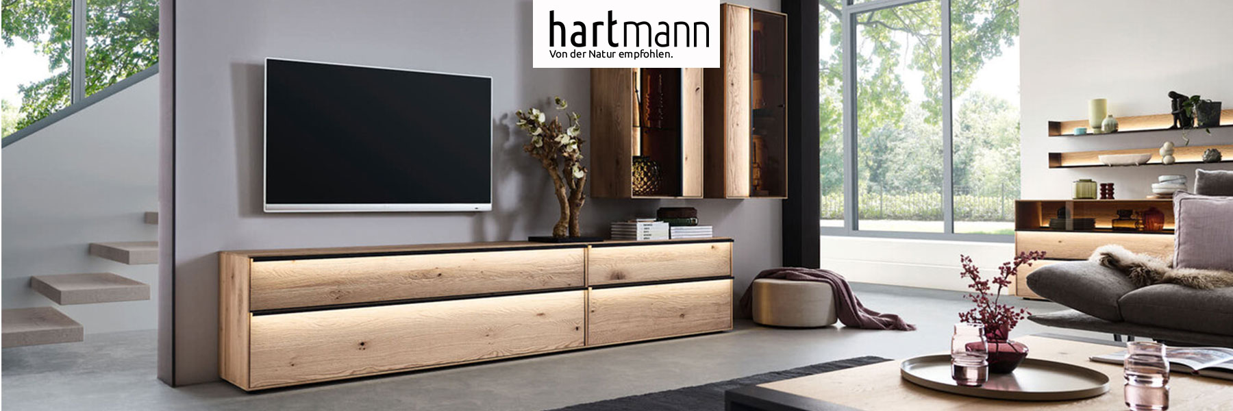 Hartmann ANEA - Massivholzmöbel aus Kerneiche Natur sandgestrahlt kombiniert mit Metall anthrazit. Ambientebild zeigt zwei schlichte Lowboards mit langen, beleuchteten Metallgriffen, darüber hängend zwei edle Vitrinen mit beleuchteter Riffholzrückwand.