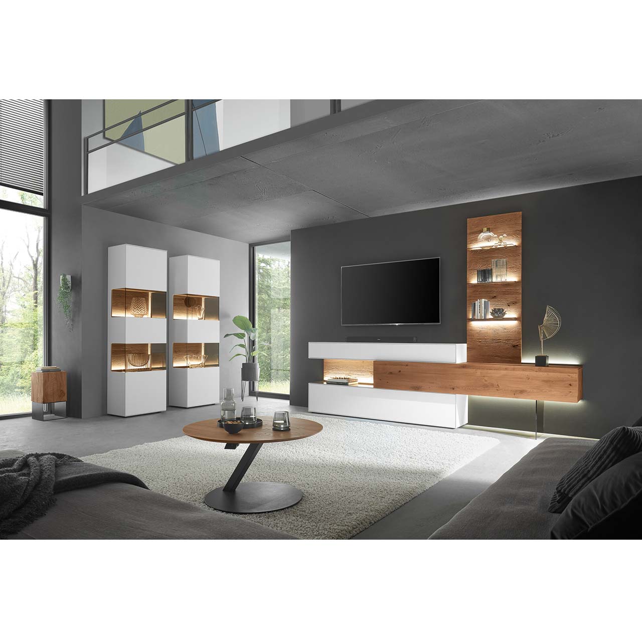 Wohnzimmer mit moderner Wohnwand aus weiß lackierter Birke und dunkler Kerneiche, bestehend aus Hängelowboard, Ober- und Unterschrank mit Beleuchtung sowie Wandpaneel mit Glasboden. Links zwei weiße Standvitrinen, davor ein runder Couchtisch.