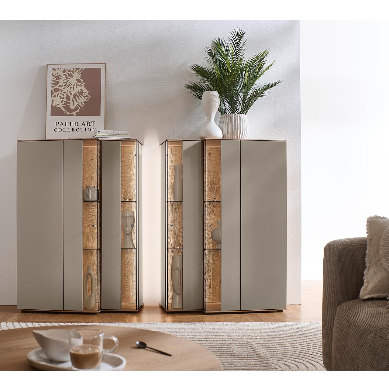 Zwei moderne Highboards aus Kerneiche Massivholz mit Lackfronten in Sand im asymmetrischen Design. Mit jeweils zwei Glas-Lacktüren, Glasböden und edler, beleuchteter Riffholzrückwand - an hellgrauer Wand stehend.