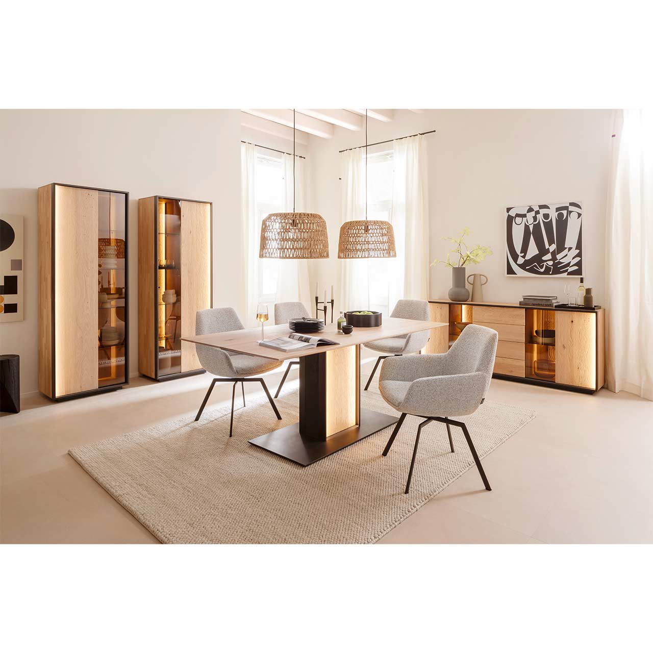 Modern eingerichtetes Esszimmer mit Massivholzmöbeln aus Kerneiche Natur mit einem rechteckigen Holztisch und vier hellen Stühlen. Rechts an der Wand ein Sideboard mit Glasausschnitt, links zwei Standvitrinen jeweils mit einer Holz-/Glastür.