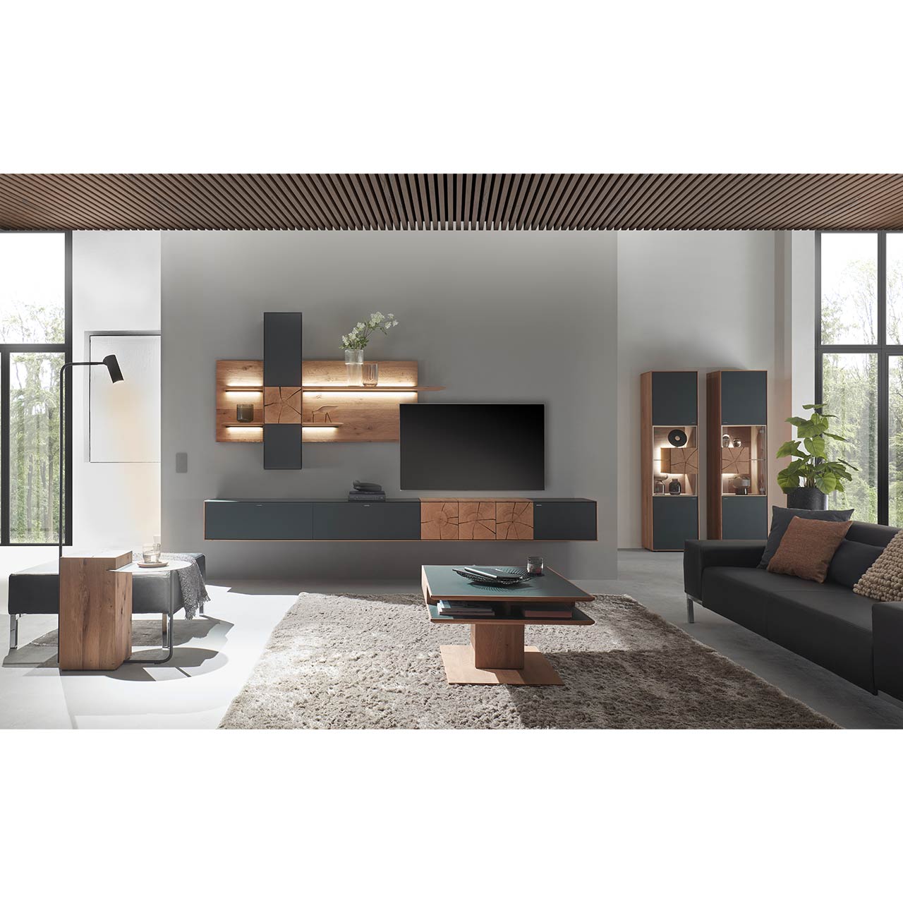 Moderne Wohnwand aus massiver Kerneiche mit anthrazitfarbenen Fronten, mit Hängelowboard, Wandpaneel mit Regalböden und Hängeschrank. Rechts im Hintergrund zwei passende Standvitrinen. Im Vordergrund ein Holz-Couchtisch mit Ablage.