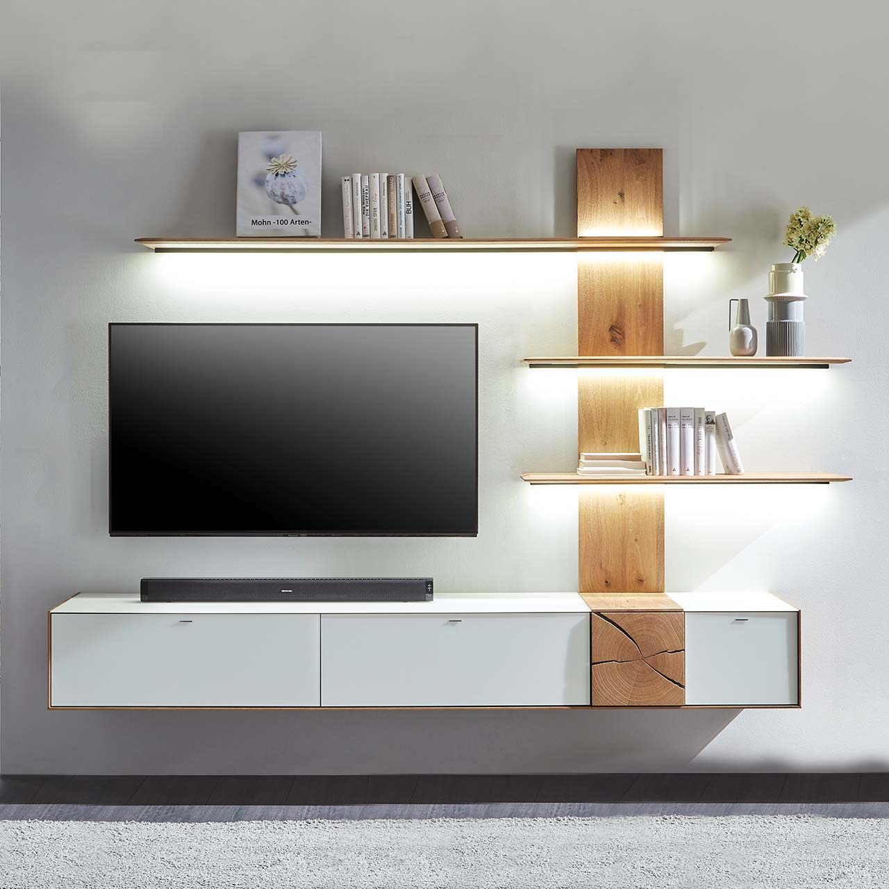 Moderne zweiteilige Massivholz Wohnkombination aus Kerneiche Umato mit Mattglas weiß Fronten und Hirnholzscheiben, mit einem langen Hängelowboard und einem Wandpaneel mit drei beleuchteten Regalböden. Dazwischen hängender Fernseher.