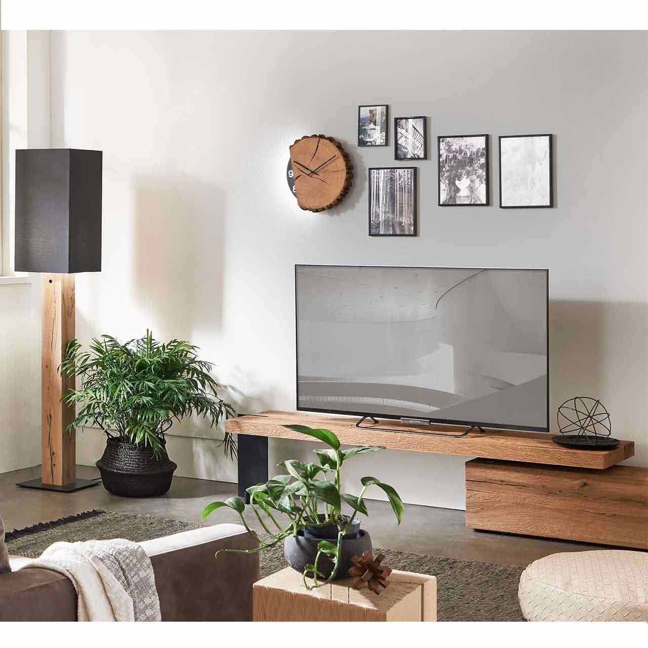Wohnzimmer mit Lowboard und Fernseher. Links daneben eine Stehlampe mit Stofflampenschirm und Holzfuß darüber hängend die passende Holzwanduhr - an grauer Wand. 