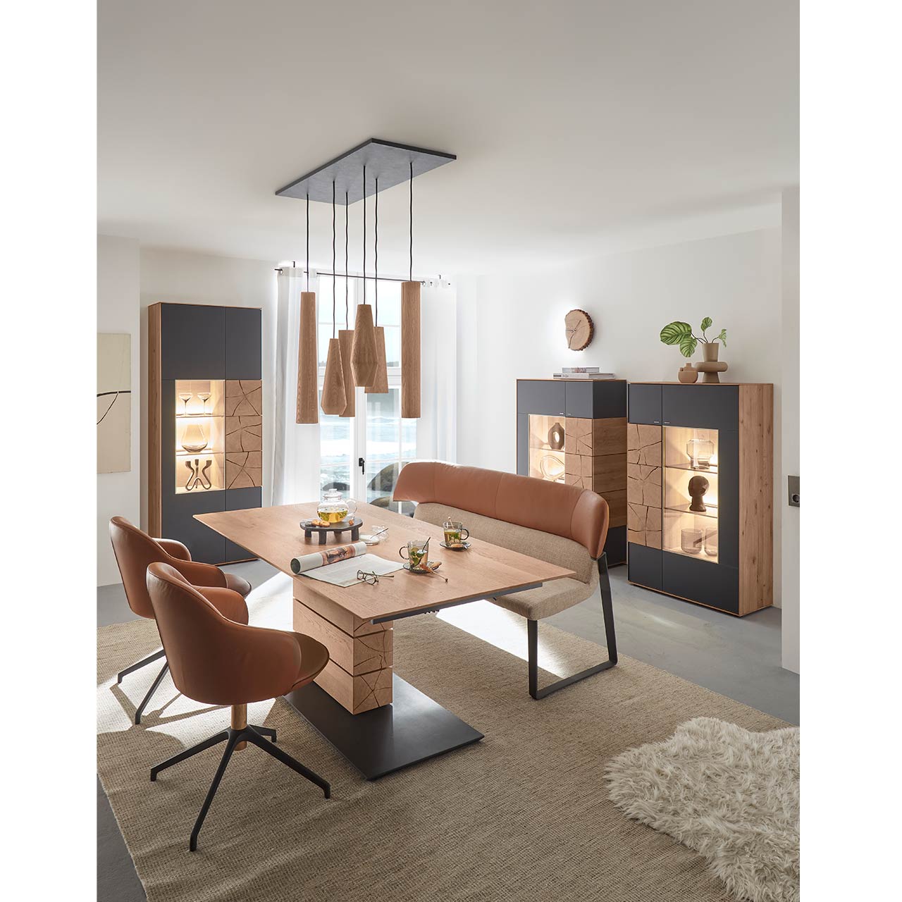 Modernes Esszimmer mit rechteckigem Massivholz Tisch mit Sitzbank und zwei Stühlen mit Lederbezug in cognac. Über dem Tisch hängt ein Pendelleuchte mit Holz-Lampenschirmen. Im Hintergrund stehen zwei passende Highboards und eine Standvitrine in Anthrazit.