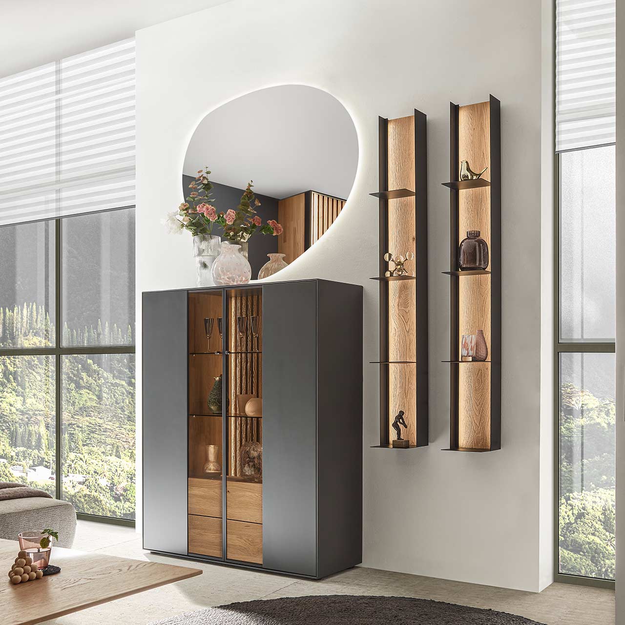 Modernes Massivholz-Highboard mit zwei Glas,- Holztüren in anthrazit, heller Holz-Lamellenrückwand und zwei Schubkästen. Darüber hängend ein asymmetrischer Wandspiegel. Rechts daneben zwei passende Metallregale - vor grauer Wand.