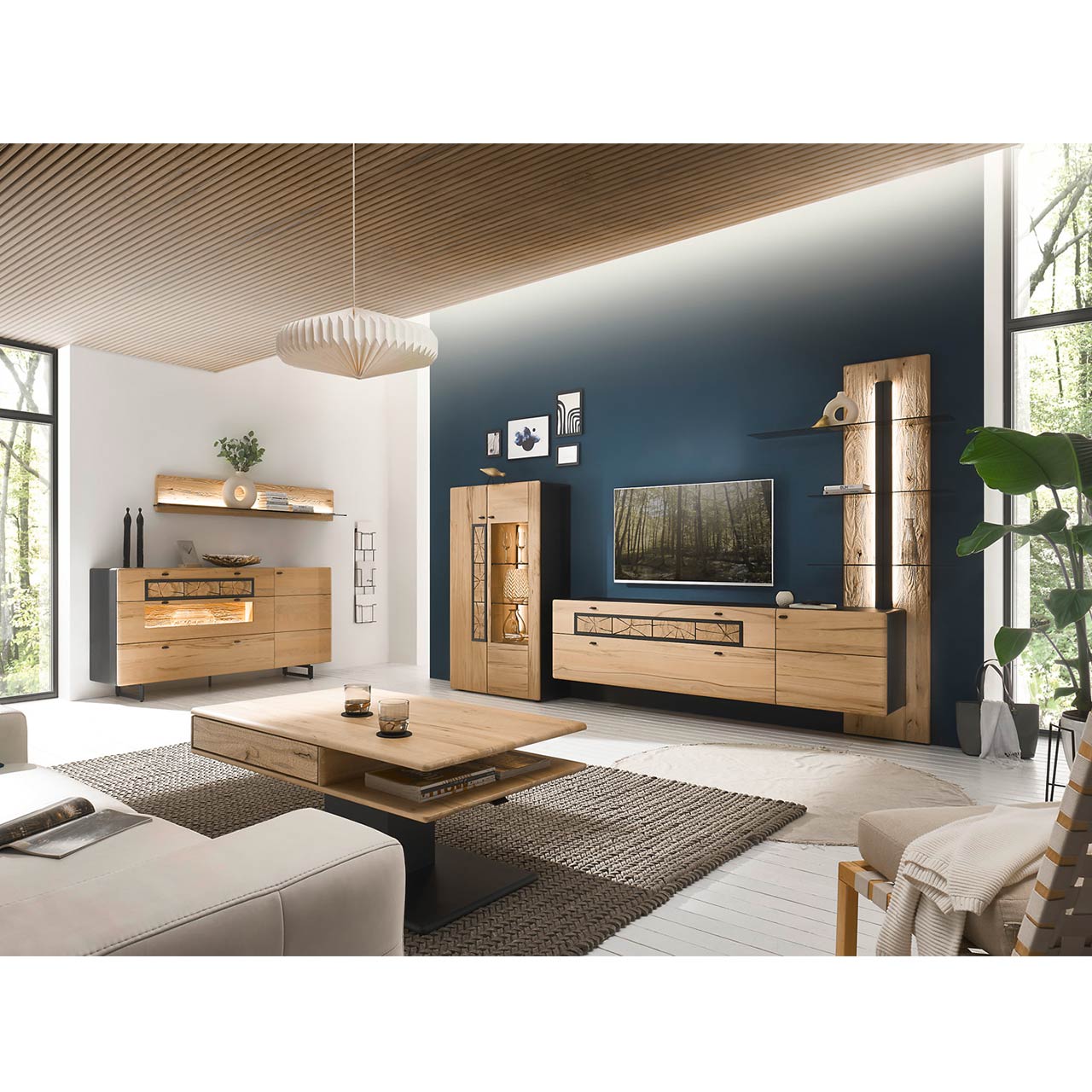 Eingerichtetes Wohnzimmer mit moderner Wohnwand aus Kernbuche vor einer dunkelblauen Wand bestehend aus Highboard, Lowboard und Paneel. Rechts das passende Sideboard auf Metallkufen darüber ein Regal. Im Vordergrund der dazugehörige Couchtisch.