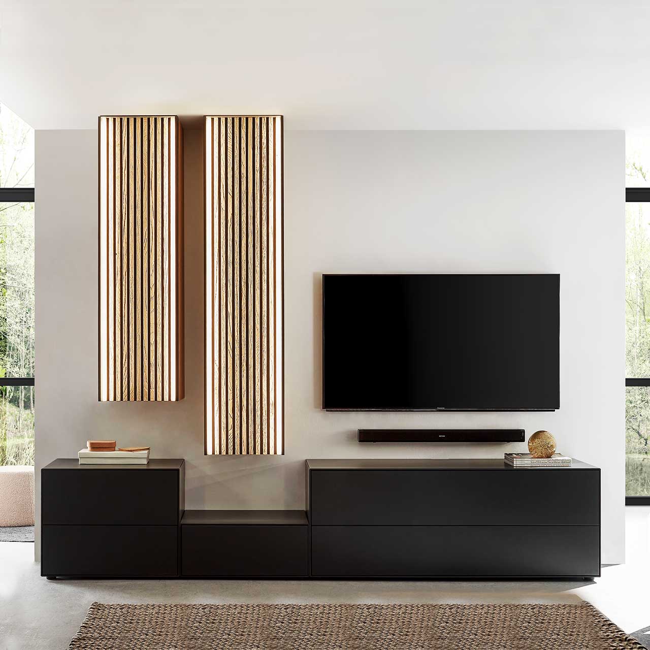 Wohnzimmer mit Fernseher und moderner fünfteiliger Wohnwand aus massivem Holz: Drei schlichte unterschiedlich hohe Lowboards anthrazit lackiert und zwei unterschiedlich hohe Hängeschränke mit hellen Holzlamellen – vor heller Wand. 
