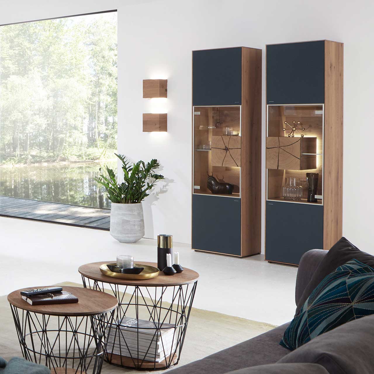 Wohnzimmerwand mit zwei modernen Standvitrinen aus massiver Kerneiche Umato mit Mattglas anthrazit Front, mittig mit beleuchtetem Glasausschnitt, Glasboden und Hirnholzblock. Links zwei Wandleuchten aus hellem Holz, im Vordergrund zwei runde Couchtische.