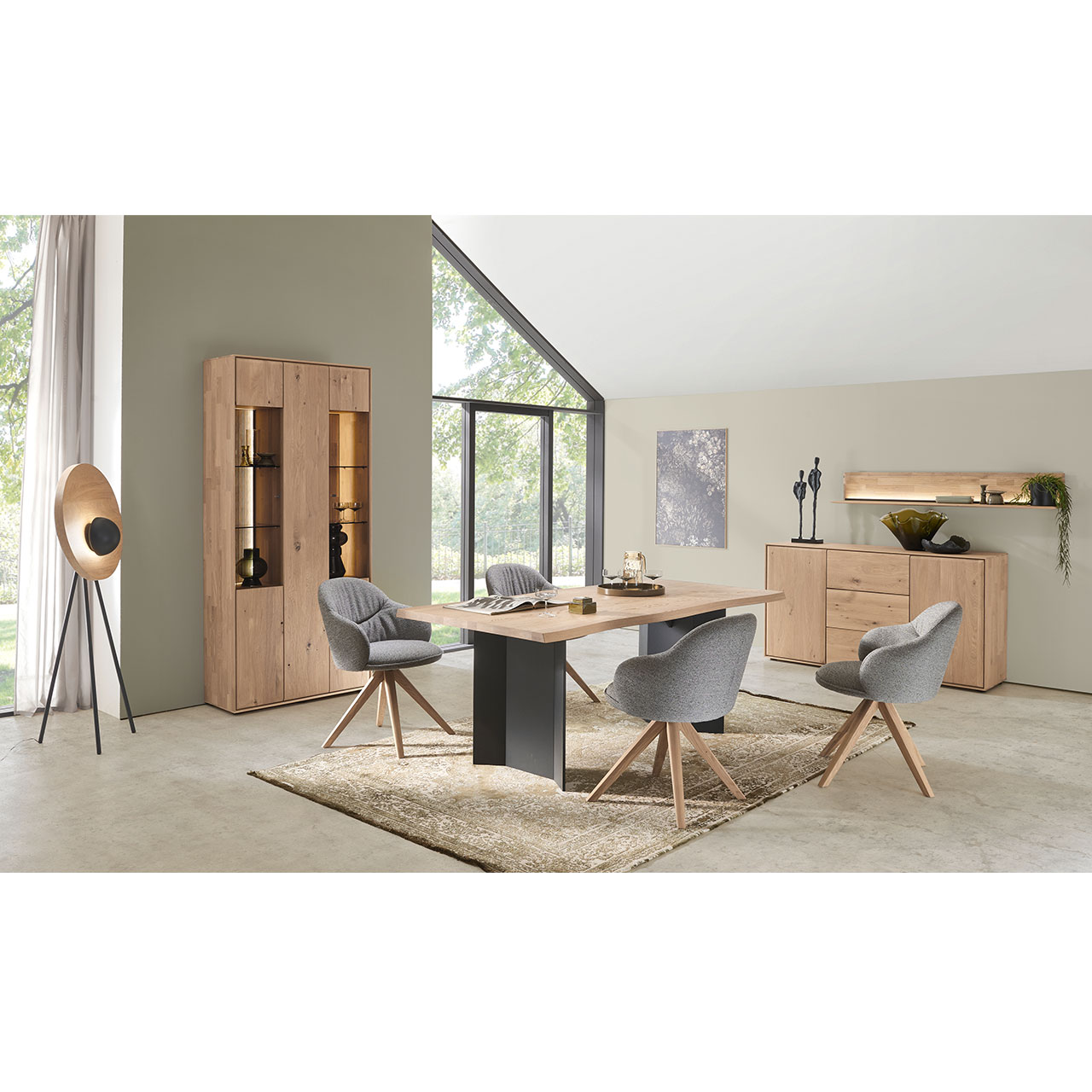 Modernes Esszimmer mit edlem Holztisch aus massiver Kerneiche mit Baumkantenprofil und vier komfortablen Stühlen mit grauem Stoffbezug und Vier-Fuß Holzgestell in moderner Stativform. Dahinter steht das passende Sideboard, links die passende Standvitrine.