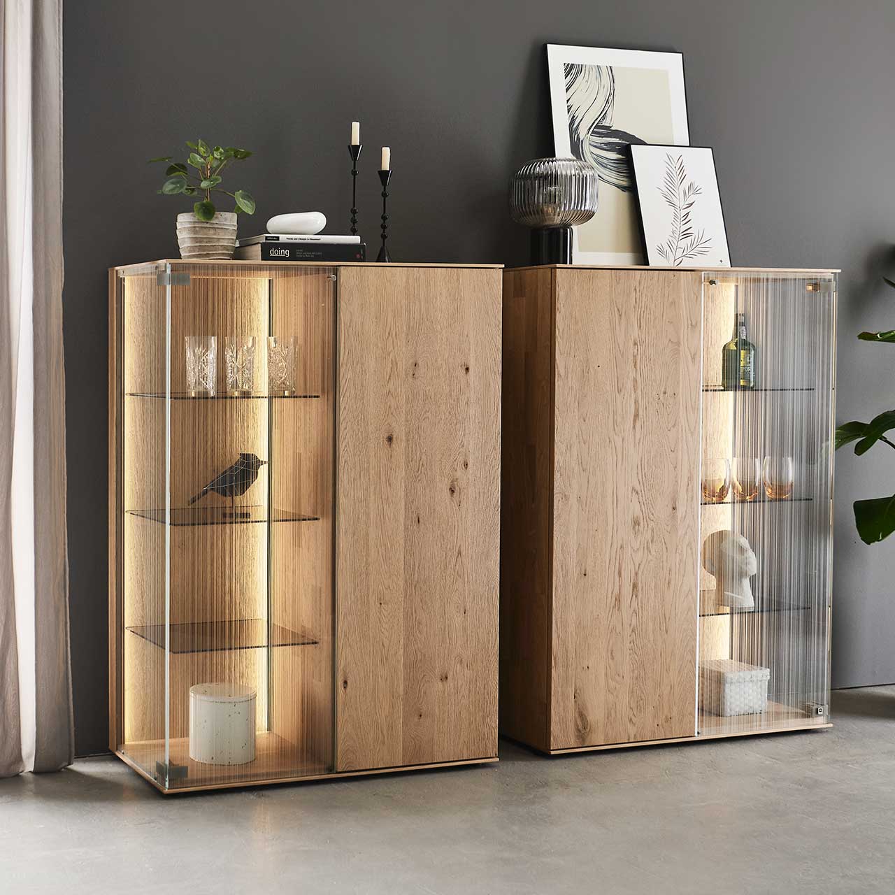 Zwei moderne Highboards aus Kerneiche Natur mit je einer Holztür und einer satinierten Glastür, drei beleuchteten Glasböden und Dekoration aus Vasen und Gläsern – vor dunkelgrauer Wand