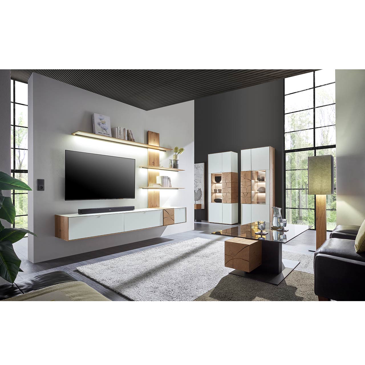 Wohnzimmer mit TV und moderner Wohnwand aus massiver Kerneiche Umato mit  Mattglas weiß Fronten und Hirnholzscheiben, mit Hängelowboard und Wandpaneel mit drei Regalböden. Rechts zwei passende Standvitrinen. Im Vordergrund Couchtisch mit Glasplatte.