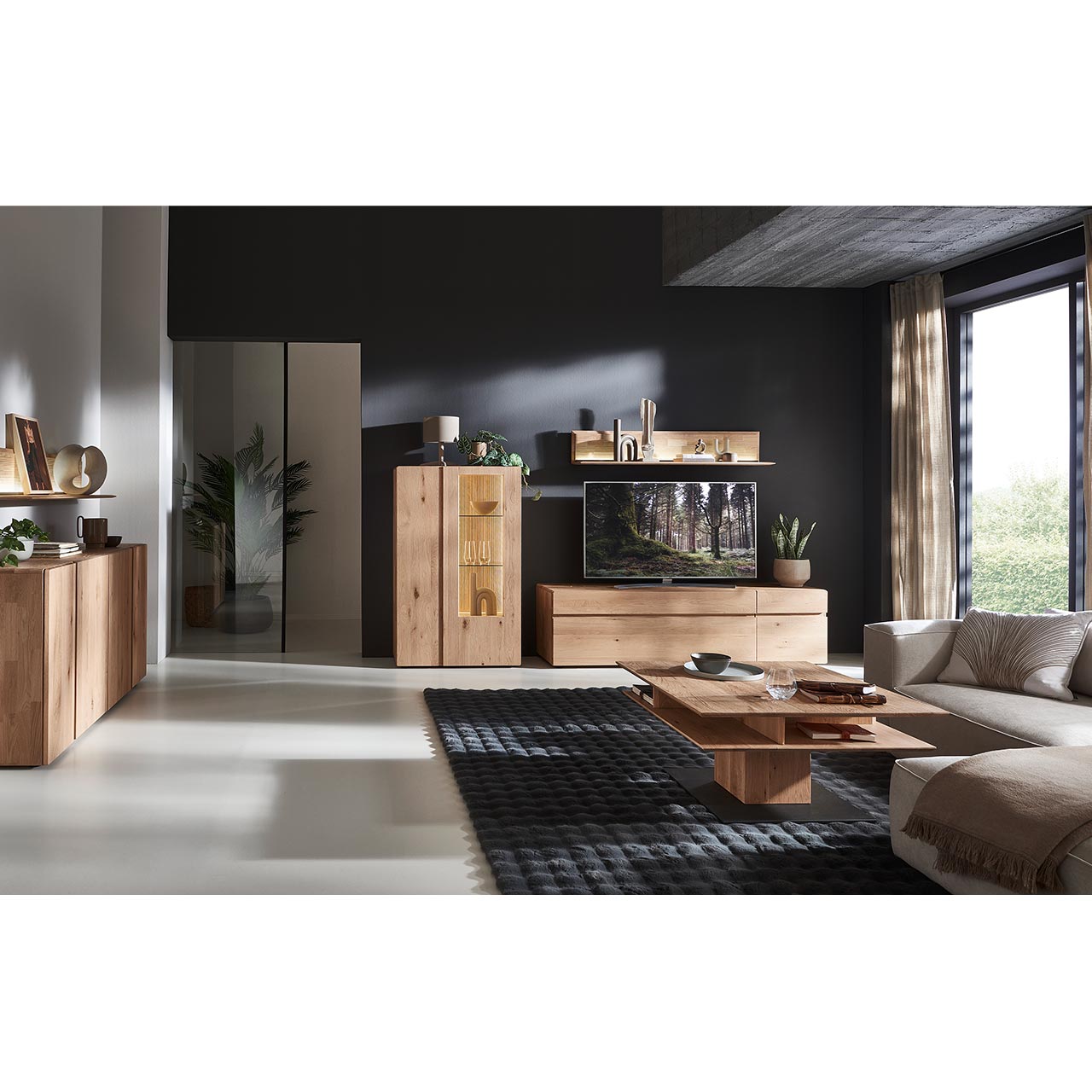 Modernes Wohnzimmer mit dreiteiliger Wohnwand aus massiver Kerneiche Natur: Lowboard, darüber hängend ein Wandpaneel mit Holzablageboden und geschroppter Rückwand. Links Highboard mit Holz-/Glastüren. Davor Holzcouchtisch, links Sideboard.