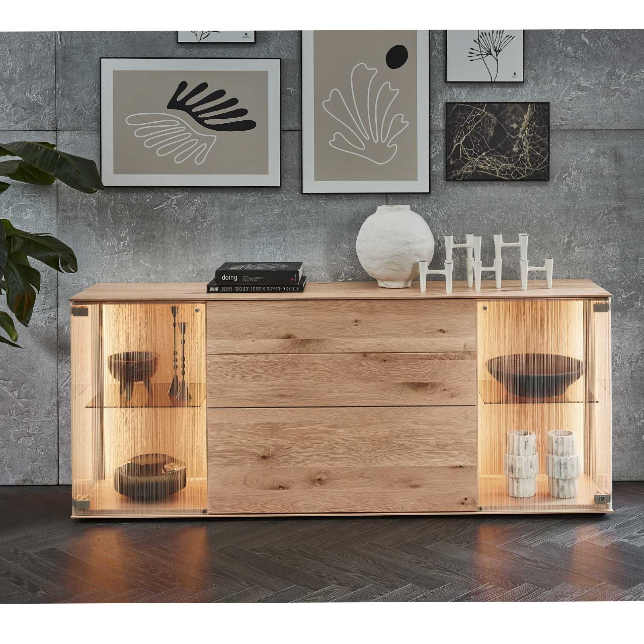 Milieubild eines Sideboards aus massiver Kerneiche Natur mit zwei beleuchteten Glastüren aus satiniertem Glas, einer Klappe und einem Schubkasten – vor grauer Wand mit Wandbildern