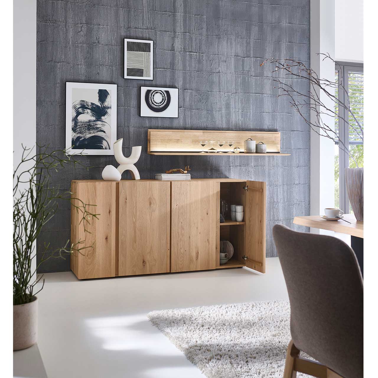 Modernes Sideboard aus massivem Eichenholz mit vier Türen. Die Tür rechts ist geöffnet und zeigt den innen liegenden Holzeinlegeboden. Über dem Sideboard hängt ein langes, beleuchtetes Wandboard.