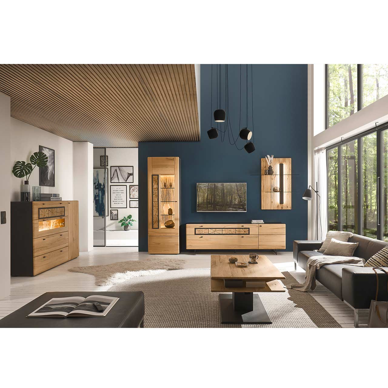 Eingerichtetes Wohnzimmer mit dreiteiliger Wohnwand aus Kernbuche vor dunkelblauer Wand: Standvitrine, Lowboard auf Metallkufen und Wandpaneel. Links das passende Sideboard mit Glasfront. Im Vordergrund der dazugehörige Couchtisch neben grauer Couch.