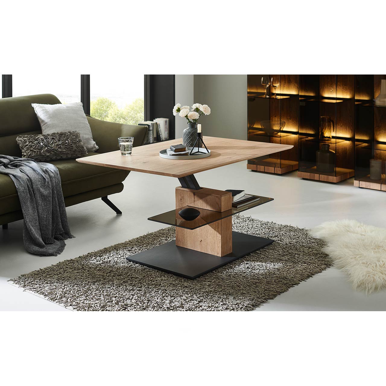 Milieubild Wohnzimmer - Hartmann NATURWERKE - Couchtisch 8400C-1424 - hochgefahren Im Wohnzimmer stehender Couchtisch mit rechteckiger, hochgefahrener Tischplatte aus Holz und zusätzlicher fester Glasablage darunter - auf massiver Holzsäule und dunkler Bodenplatte.