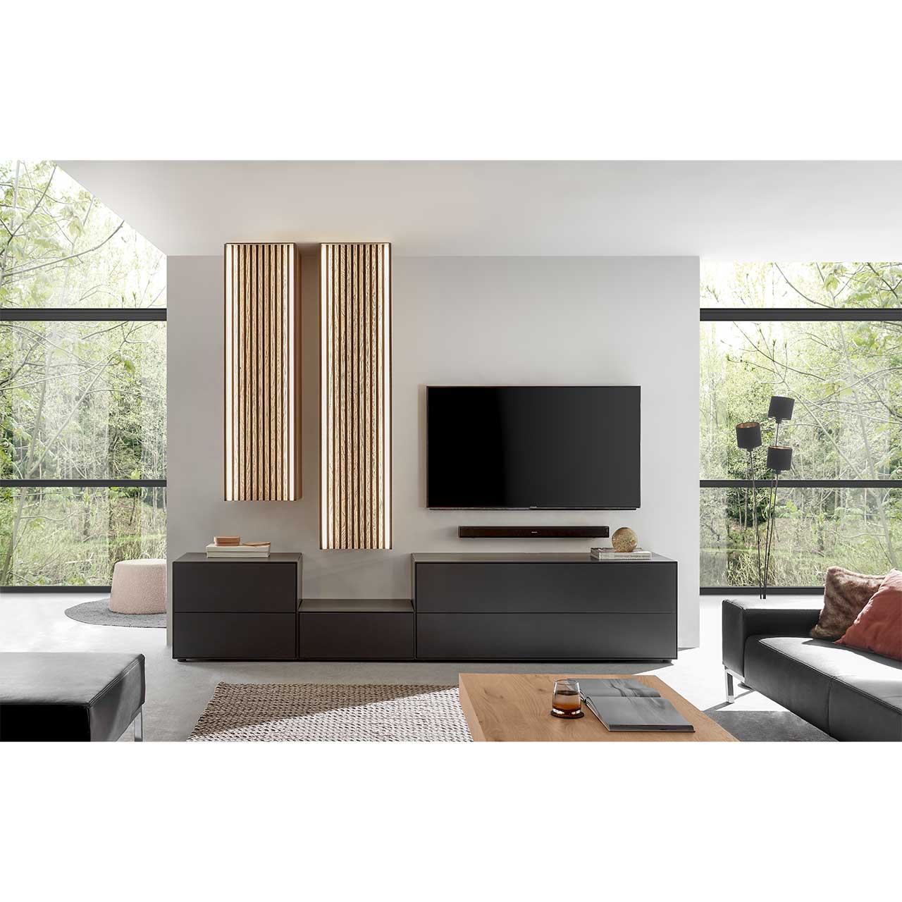 Wohnzimmer mit Fernseher und moderner fünfteiliger Wohnwand aus massivem Holz: Drei schlichte unterschiedlich hohe Lowboards anthrazit lackiert und zwei unterschiedlich hohe Hängeschränke mit hellen Holzlamellen – vor heller Wand. 