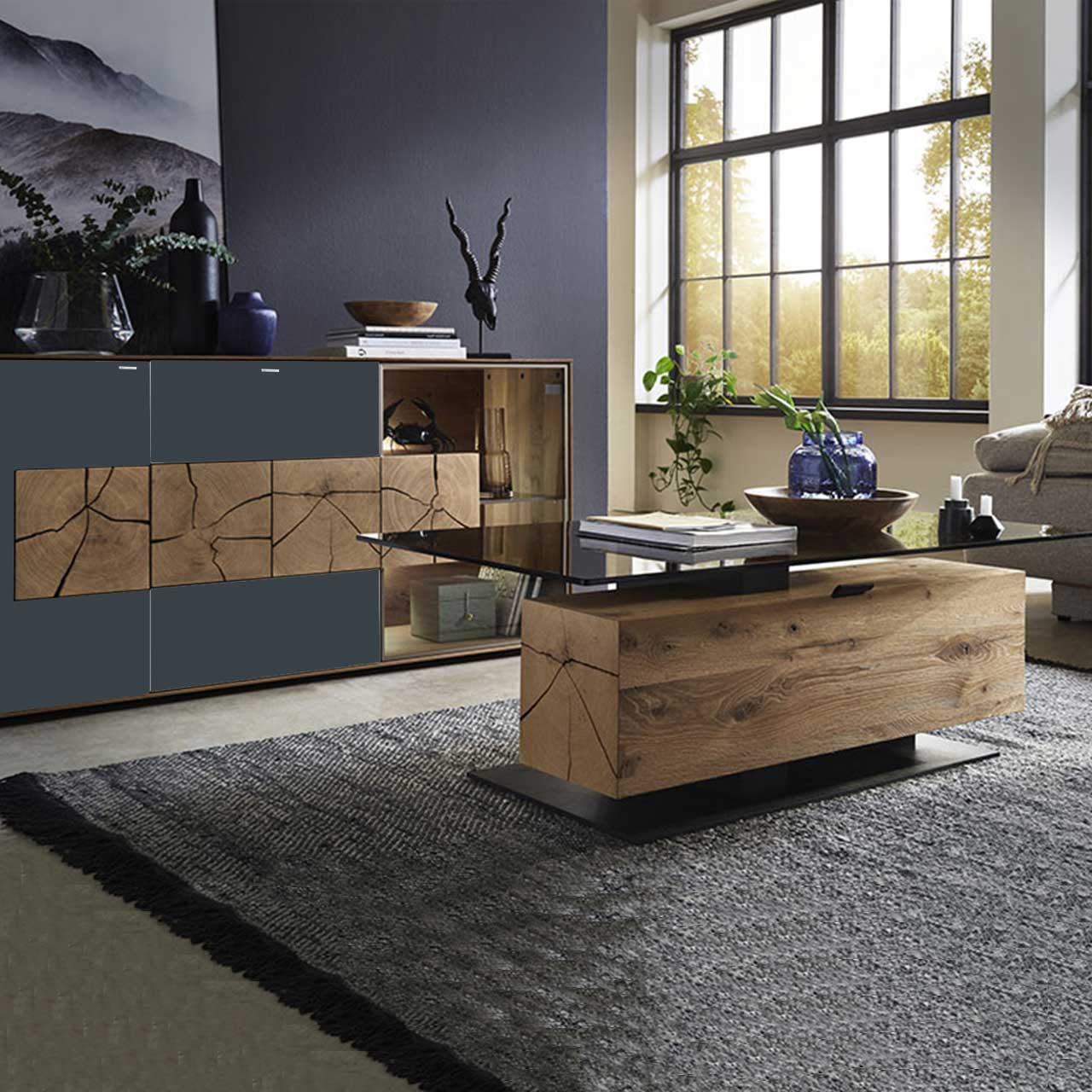 Bildausschnitt eines Wohnzimmers mit modernem Lowboard aus massiver Kerneiche Umato mit anthrazitfarbenen Mattglasfronten, drei rechteckigen Hirnholzscheiben und Glastür mit beleuchtetem Glasboden und Hirnholzblock, davor passender Couchtisch.