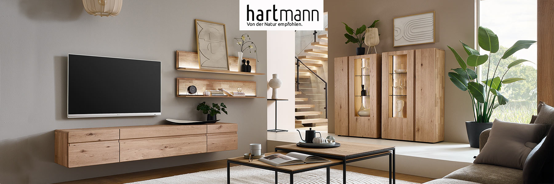 Hartmann VIDA - Massivholzmöbel aus Kerneiche Natur kombiniert mit geschoppten Rückwänden und Holzeinkerbungen. Ambientebild zeigt Hängelowboard, zwei beleuchtete Wandregale mit Holzablageböden und zwei passende Highboards mit Holz-/Glastüren.