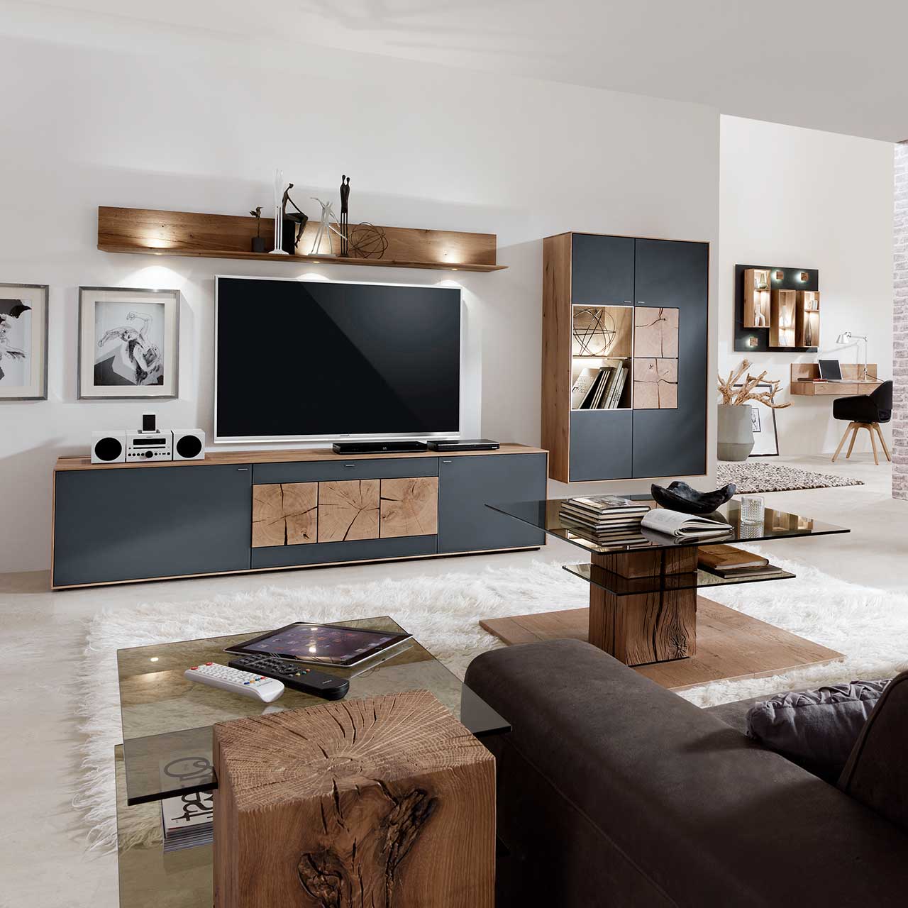 Wohnzimmer mit TV und moderner Wohnwand aus massiver Kerneiche Umato mit Mattglas anthrazit Fronten: Langes Lowboard mit dekorativen Hirnholzscheiben, Hängeregal aus Kerneiche, Hängevitrine mit beleuchtetem Glasausschnitt und Hirnholz.