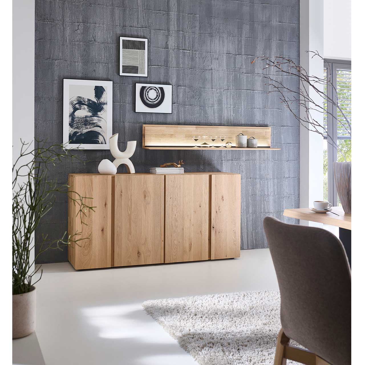 Milieubild - Hartmann VIDA - Sideboard 8630-5191 - Kerneiche Natur Cleanes Sideboard aus massivem Eichenholz mit vier Türen und zwei markanten Holzeinkerbungen in den Fronten - im Wohnzimmer stehend. Darüber hängt ein langes beleuchtetes Wandboard.