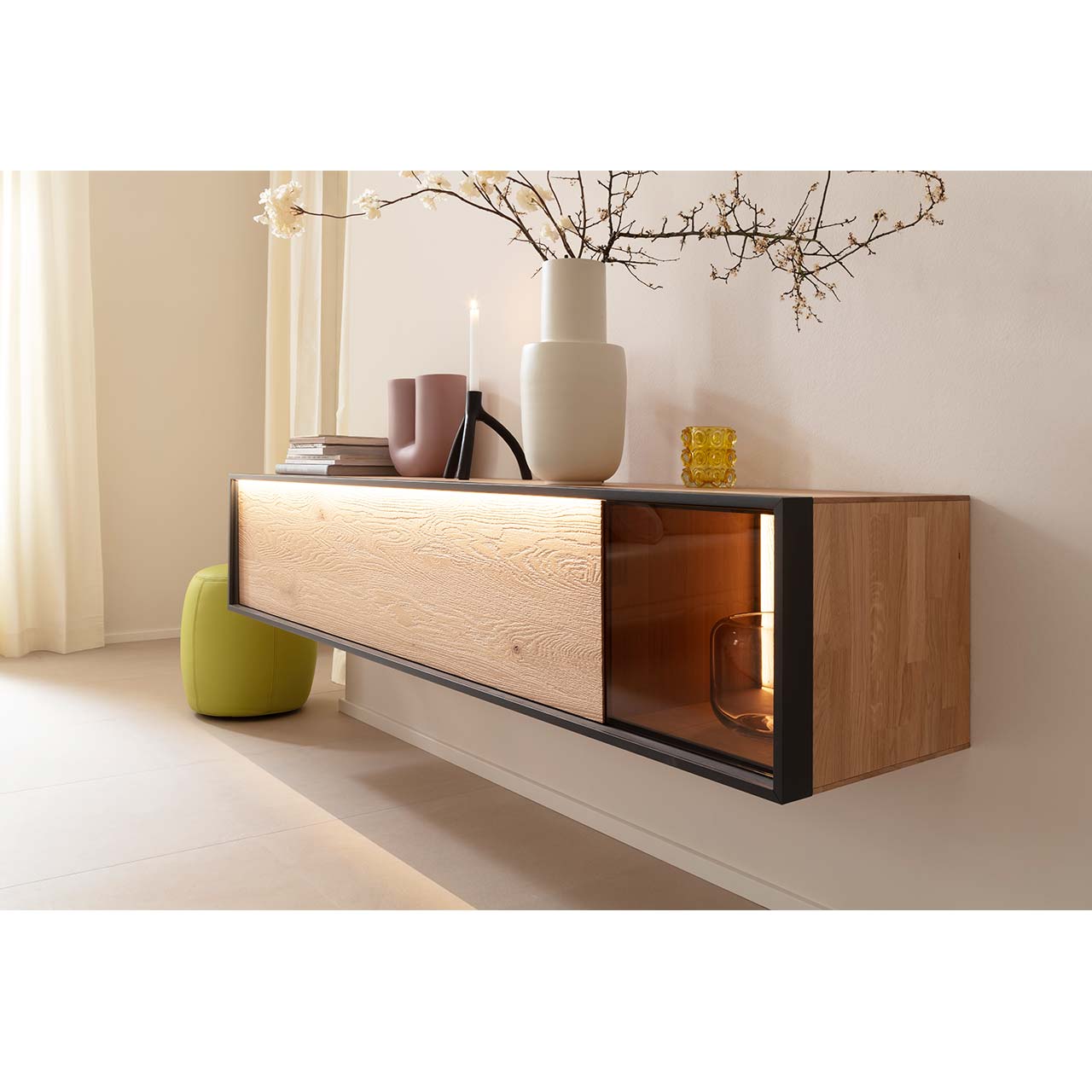 Modernes Hängelowboard aus sandgestrahlter Kerneiche Natur mit beleuchtetem Glasausschnitt rechts, eingefasst in einen anthrazitfarbenen Metallrahmen - an beiger Wand hängend.