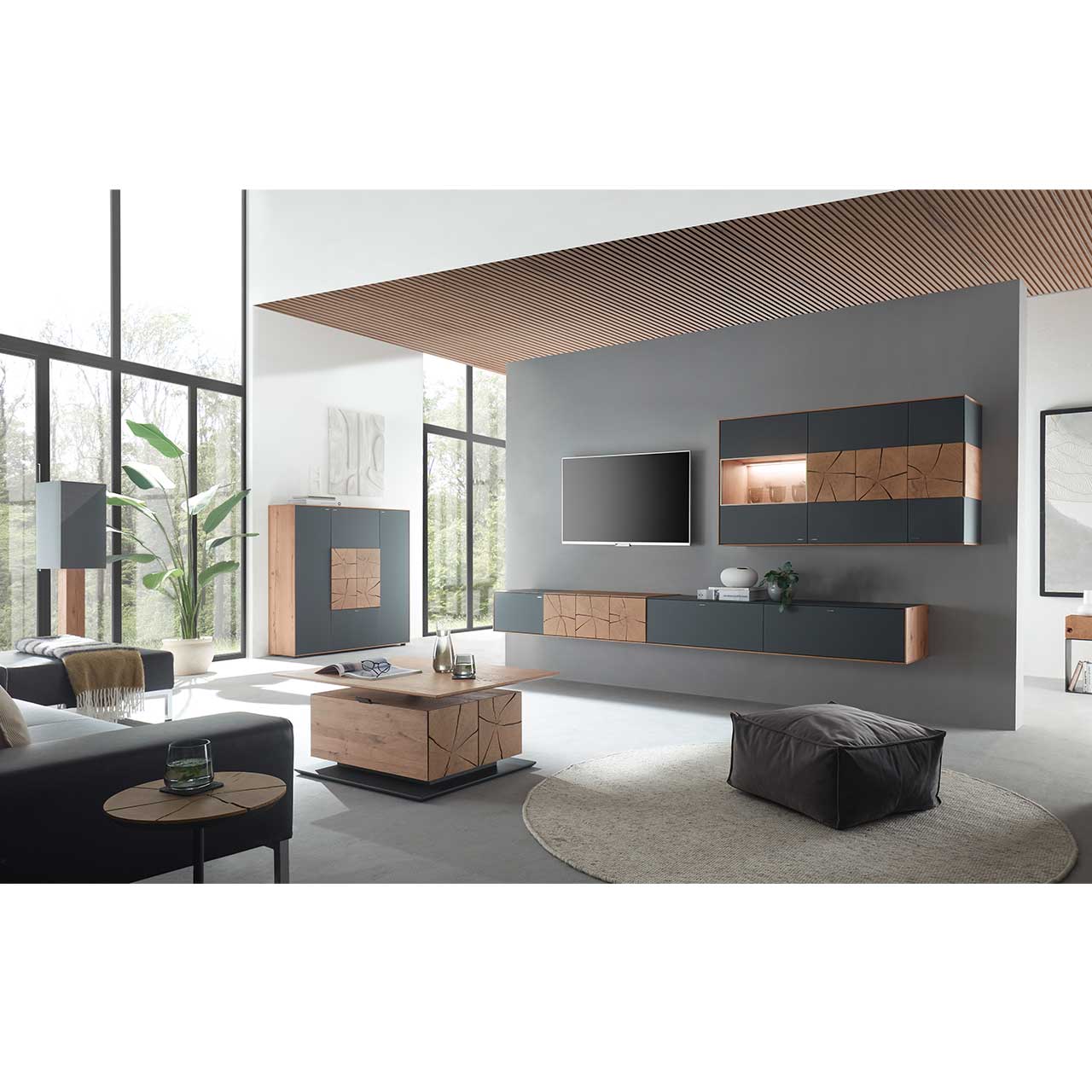 Wohnzimmer mit TV und moderner Wohnwand aus massiver Kerneiche und Mattglas anthrazit Fronten, mit schmalem Hängelowboard und beleuchteter Hängevitrine. Links das passende Highboard. Im Vordergrund ein rechteckiger Holz-Couchtisch.
