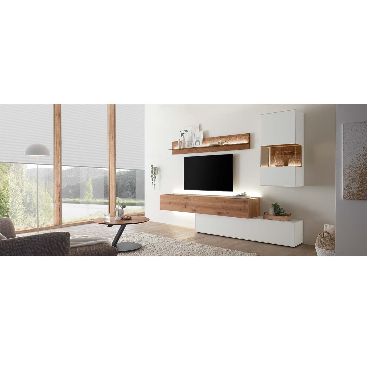 Wohnzimmer mit moderner Wohnwand aus weiß lackierter Birke und dunkler Kerneiche Umato bestehend aus Hängelowboard, einem weißen Unterschrank, einer weißen Hängevitrine und einem Wandregal mit Glasablageboden. Davor ein runder Couchtisch.