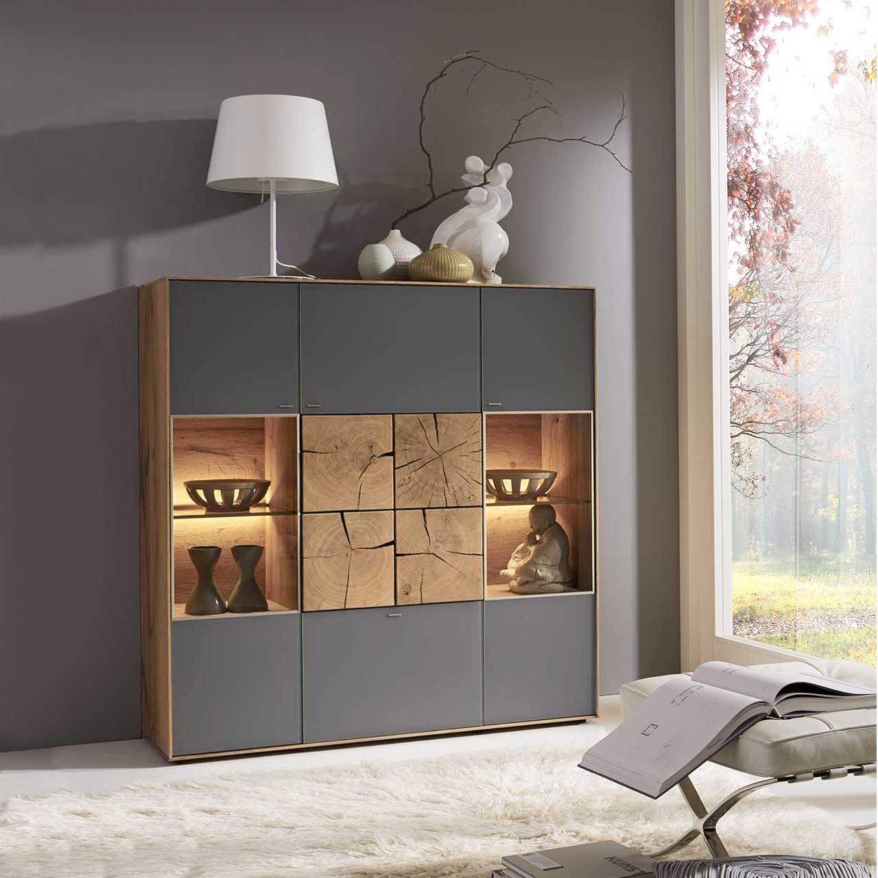 Modernes Highboard aus massiver Kerneiche Umato mit Mattglas anthrazit Fronten, rechts und links mit beleuchtetem Glasausschnitt, mittig mit vier dekorativen, rechteckigen Hirnholzscheiben in heller Kerneiche Umato - in einem modernen Flur stehend.